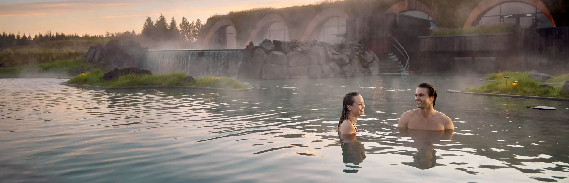 Two people bathing at Laugarás Lagoon with steam rising from the water.
