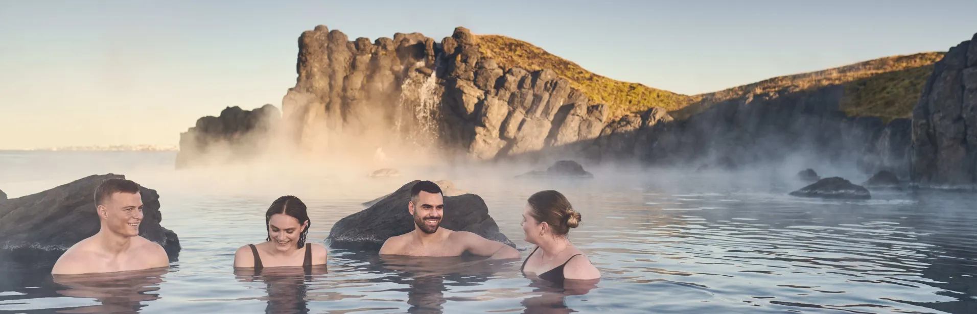 Four people bathing at Sky Lagoon among rocks and rising steam.